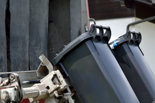 The image shows a close-up of two black plastic rubbish bins attached to a metal frame, positioned on a driveway or pavement area. The bins are designed with hinged lids, one of which is slightly open, revealing a glimpse of the interior. The surface of the bins appears smooth and slightly reflective, with a matte finish. They are secured to a metallic towing or support apparatus, which is part of a waste collection vehicle or trailer. The surrounding environment includes dark wooden panels or fencing in the background, indicating an outdoor setting likely within a residential or commercial area in Hampstead. The lighting is natural, suggesting daytime, and the scene emphasizes the process of rubbish collection or disposal, fitting with the services offered by Office Clearance Hampstead for efficient waste removal and management in the local area.