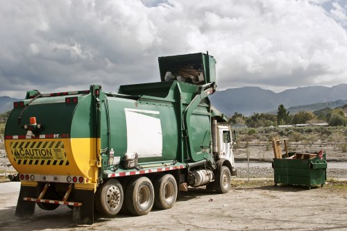 A large green waste collection truck with a yellow caution marking on its rear is parked on an open gravel area, with its side compactor raised, ready for unloading. The truck has a white cab and visible black tires, positioned on the right side of the image. To the right of the truck, there is a green waste container filled with various debris, sitting on the ground near a weathered metal fence. The background features a scenic view of distant hills or mountains under a partly cloudy sky, suggesting an outdoor waste management or rubbish removal operation that might be typical in or near Hampstead or surrounding areas. The environment appears rural or on the outskirts of an urban area, with natural landscape elements visible beyond the fence. This scene exemplifies professional rubbish collection services, such as those provided by Office Clearance Hampstead, and highlights equipment used in waste removal services in the local area.