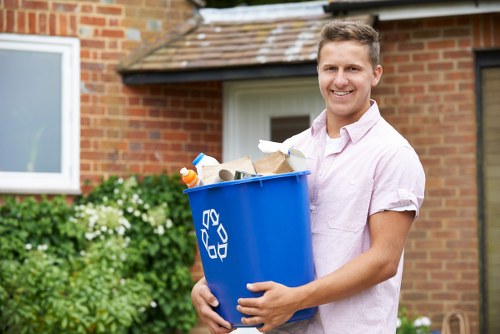 A young man with short, light brown hair and a friendly smile is standing outdoors in front of a red brick building with a white window, holding a large blue recycling bin filled with various household waste items, including paper, cardboard, and plastic bottles. He is wearing a light pink, short-sleeved shirt and appears to be preparing for rubbish removal or recycling collection. The background shows greenery, including a bush with white flowers, indicating a residential setting in Hampstead or nearby. The scene is illuminated by natural daylight, suggesting a clear and bright day, and highlights the practical aspect of rubbish collection services provided by Office Clearance Hampstead, situated in an area typical of London’s northwest suburbs, focusing on domestic and small commercial cleanouts.
