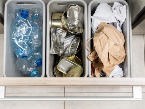 The image displays three grey plastic sorting bins filled with waste materials, positioned on a tiled floor in what appears to be an indoor or outdoor area in Hampstead. The left bin contains several clear plastic bottles with blue caps, some partially crushed, and a few smaller plastic containers. The middle bin holds a collection of used aluminum cans, including beverage cans, stacked and overlapping, with visible ridged surfaces and metallic finishes in silver and gold tones. The right bin contains crumpled paper and cardboard, including a crumpled brown paper bag and white paper sheets, with some edges sticking out above the bin's rim. The environment is illuminated with even lighting, casting minimal shadows, and the image emphasizes waste segregation, suitable for a professional rubbish removal service such as Office Clearance Hampstead. The background features a light-colored tiled surface that complements the tidy and organized appearance of the bins, reflecting typical waste collection practices for disposal or recycling in a Hampstead office or premises context.