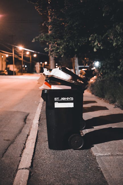 A street scene featuring a large white rubbish collection truck positioned alongside the curb, with its rear open, revealing an interior filled with various waste materials. The truck has signs of rus