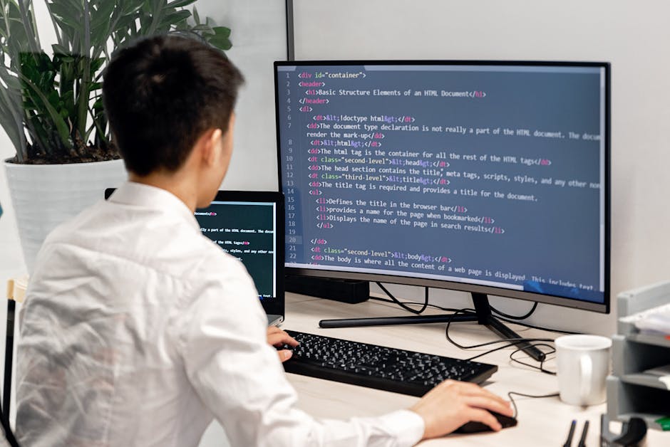 A close-up view of a computer workspace displaying lines of code on a large monitor, with a glowing blue light emanating from the screen. Behind the main monitor, there is another screen with addition