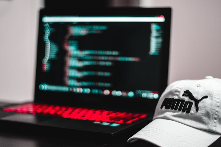 A close-up image of a black laptop with a slightly blurred screen displaying lines of colorful code, including blues, reds, and whites, in the background. In the foreground, a white Puma baseball cap