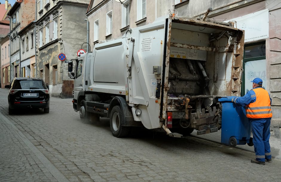 A waste collection vehicle is parked on a narrow cobblestone street amidst residential buildings, with its large rear compartment open and partly rusty, indicating it is used for rubbish collection an