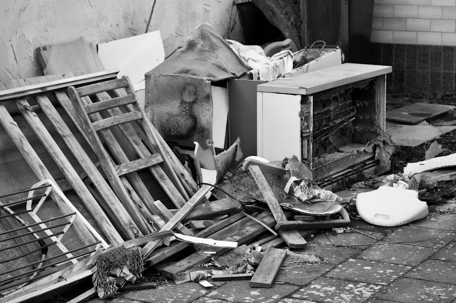 In the foreground, a pile of mixed household waste items is visible, including a large, rectangular white plastic container, dark brown cardboard boxes, and several plastic bags. The items are loosely